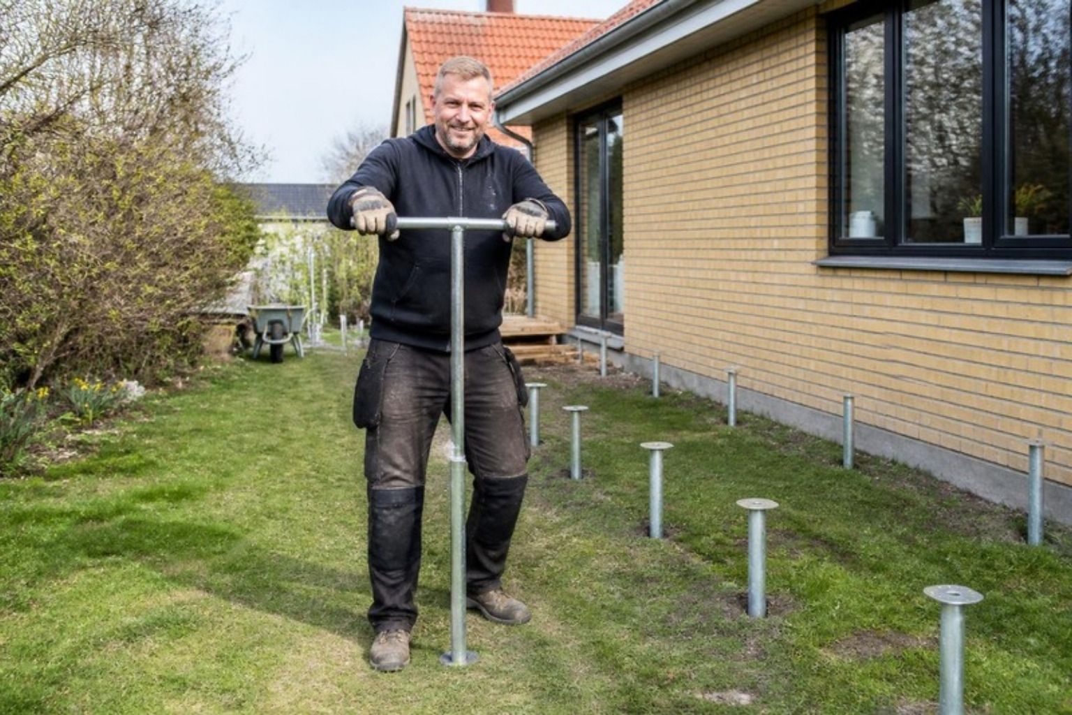 Smilende håndværker skruer en jordskrue ned i græsplænen ved et gult murstenshus, hvor flere jordskruer allerede står på række