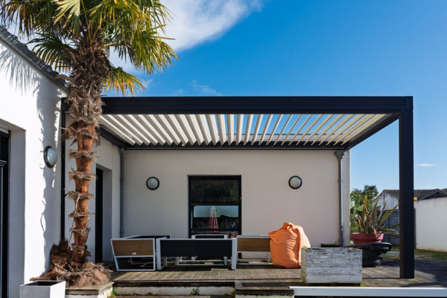 Sort lamelpergola over en træterrasse ved et hvidt hus med palme og loungemøbler