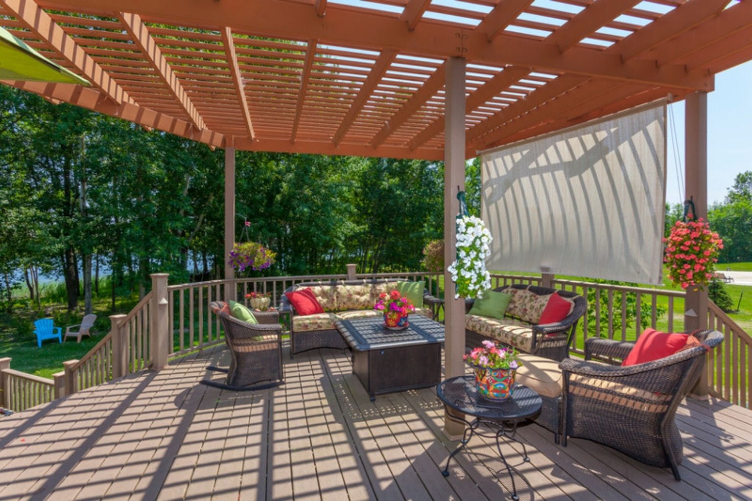 Træpergola med hvidt solsejl over en træterrasse med flettede loungemøbler og træer