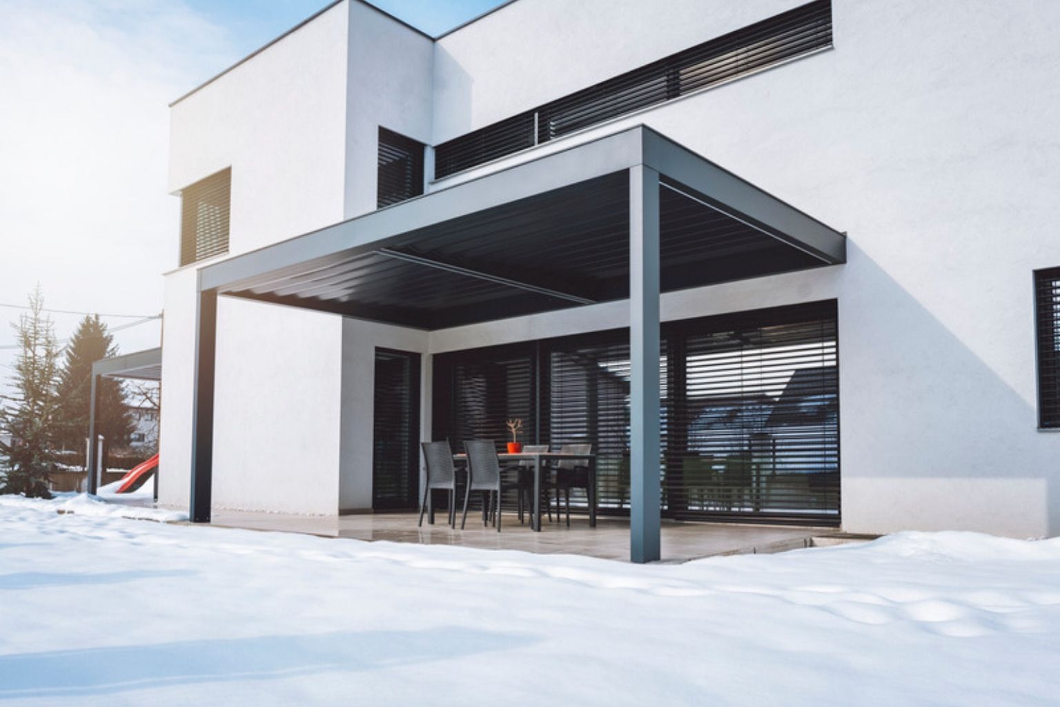 Sort lamelpergola over spisebord ved et moderne hvidt hus med sne i haven