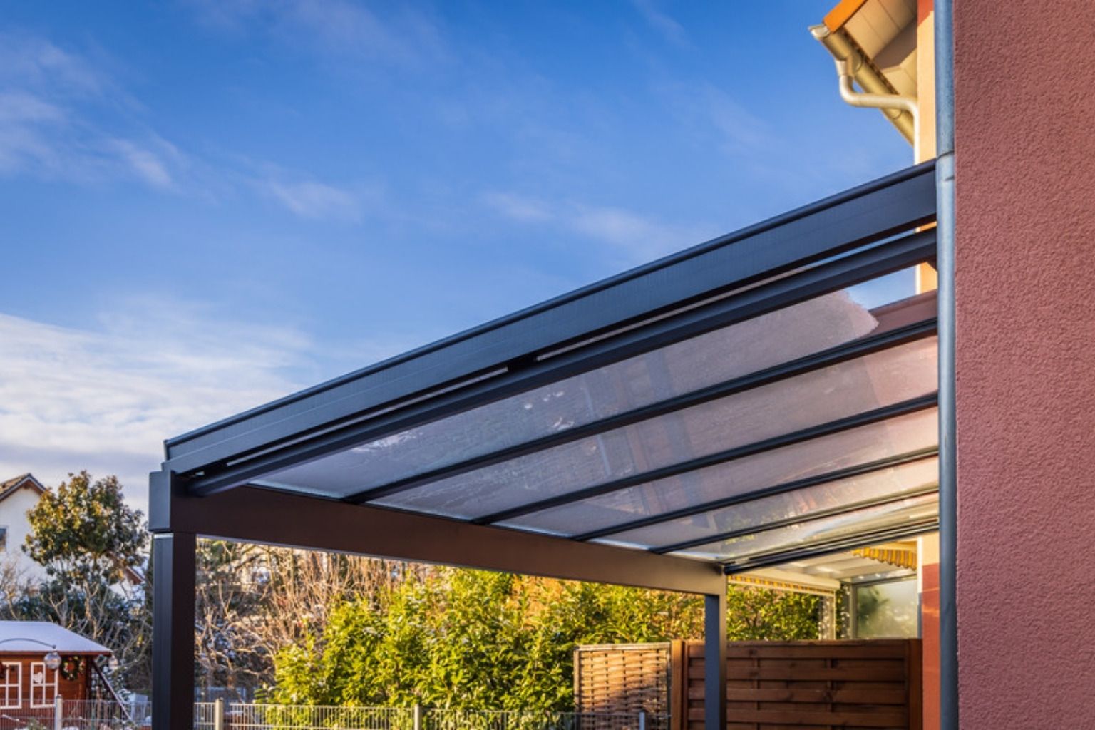 Terrasseoverdækning med sort aluminiumsramme og klare tagplader mod blå himmel og træer